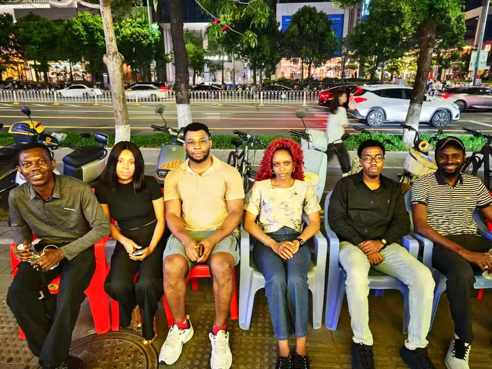 Six members of Nigerians in Shenzhen seated in a row on a Shenzhen street at night, with traffic and shopfronts behind them