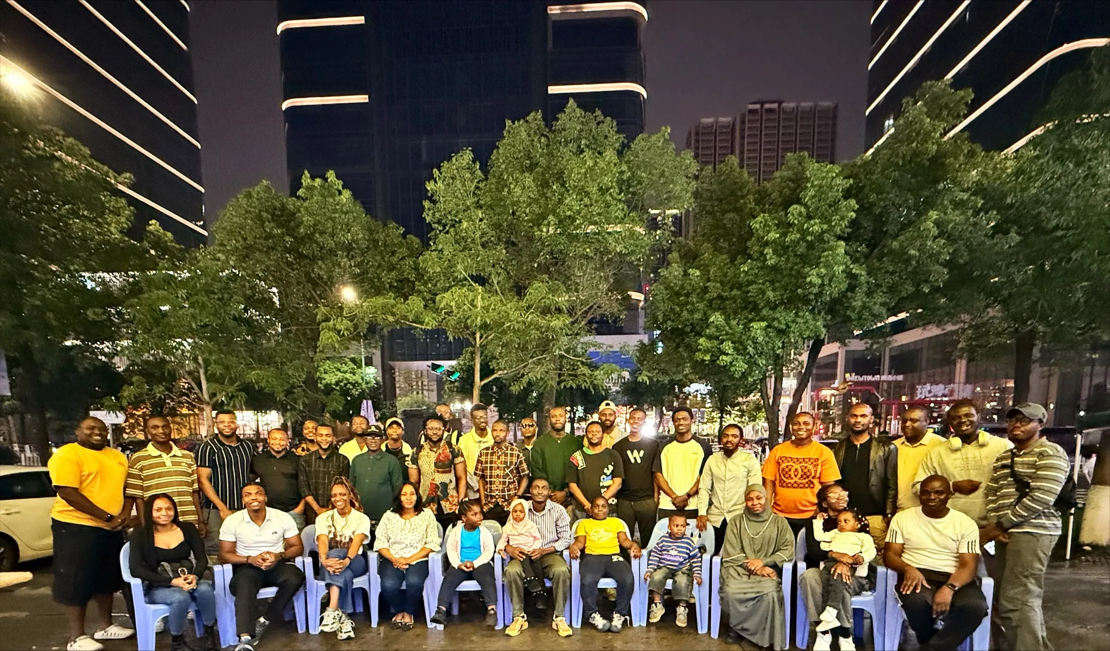 About forty members of Nigerians in Shenzhen photographed together at night on a Shenzhen street, seated on plastic chairs in front of trees with lit towers behind