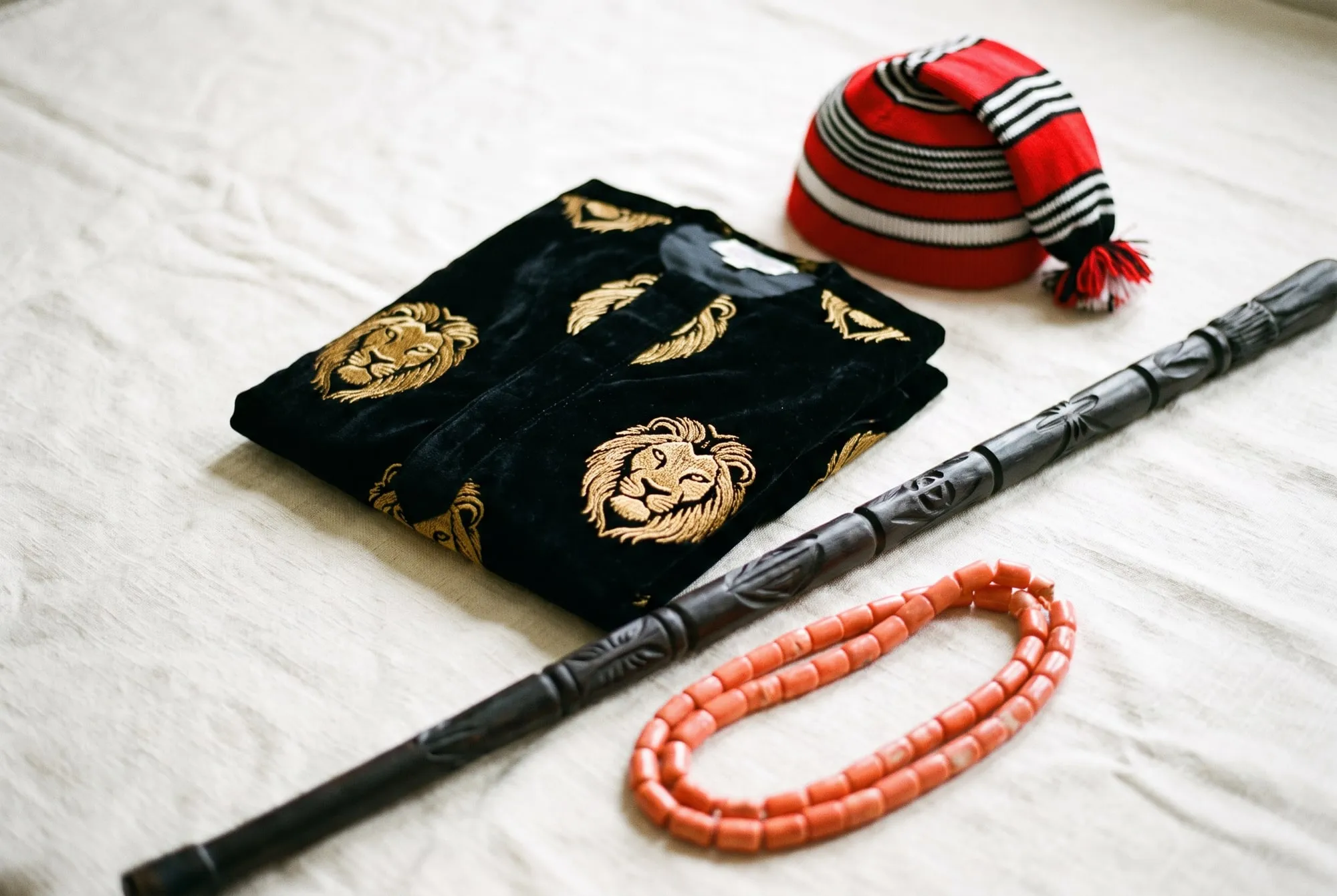 Nigerian ceremonial dress photographed as a still life: an embroidered shirt, a woven cap, a carved staff and coral beads